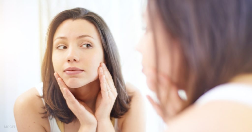 Woman (model) looks in mirror wondering what is a fair price to pay for a BOTOX treatment